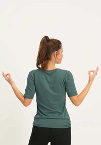 Türkises, kurzärmeliges, figurbetontes Oberteil mit rundem Halsausschnitt und glatter Textur. Das Modell ist von hinten positioniert, die Hände in einer Mudra-Position.