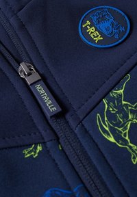 Tissu bleu marine imprimé de dinosaures, un patch T-Rex et un curseur de fermeture éclair portant l’inscription « NORTHVILLE » sur un vêtement.