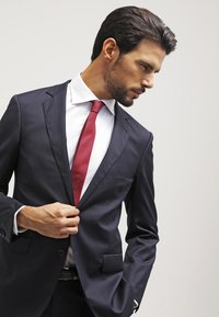 Námornícky modrá saka s jednou kapsou, biela košeľa a červená kravata s vzorom. Model si upravuje sako jednou rukou.