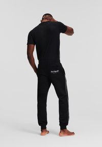 Cămașă neagră cu mânecă scurtă și pantaloni de trening negri cu dungi albe pe laterale. Pantalonii au un buzunar la spate cu detalii cu logo.