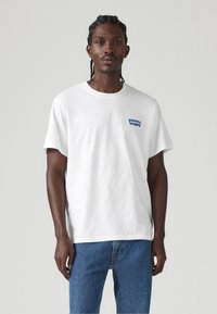 Baltas medvilnės t-Shirt su apvaliu kaklu ir trumpomis rankovėmis, dešinėje krūtinės pusėje esantis mėlynas Levi's logotipo ženklelis. Derinamas su mėlynomis džinsinėmis kelnėmis.