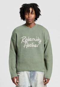 Lichtgroene oversized trui van gestructureerde stof, met een V-hals en witte bedrukte tekst "Relenity Atelier" aan de voorkant.