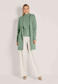 Vrouw in een lichtgroene gebreide cardigan en trui, met witte wijde pijpen broek en beige hakken, staand tegen een effen achtergrond.