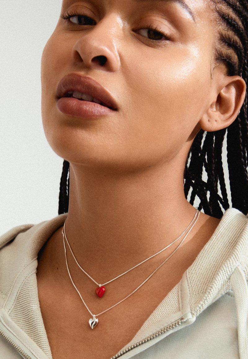 Des colliers en argent superposés avec un charme en forme de cœur rouge et un pendentif en forme de cœur en argent, portés sur une peau nue, associés à un hoodie zippé de couleur claire.
