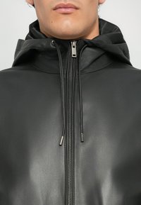 Lähivõte inimesest, kes kannab musta nahast tõmblukuga jakk, millel on kapuuts ja nöörid, näidates alalõua ja ülemise rinnaosa.