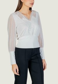 Biała bluzka z przezroczystymi, długimi rękawami w groszki oraz ozdobną koronką na przodzie, zestawiona z czarnymi, dopasowanymi spodniami.