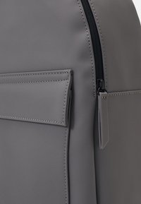 Sac à dos gris avec une texture lisse et mate, doté d'une poche avant et d'une fermeture éclair avec une petite étiquette attachée. Cousu avec soin, les points de couture sont bien visibles.
