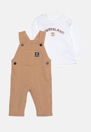 Salopette en coton marron avec des bretelles ajustables et des accents en bouton, assortie d'une chemise à manches longues blanche arborant un logo multicolore.
