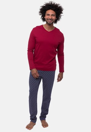 Mann mit lockigem Haar lächelt, barfuß, trägt ein rotes Langarmshirt und gemusterte dunkelblaue Pyjamahosen vor einem einfarbigen Hintergrund.