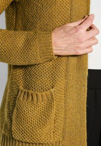 Eine Hand hält die offene Vorderseite einer senfgelben, strukturierten Strickjacke mit gerippten Kanten und kleiner Vordertasche über einem weißen Oberteil und schwarzen Hosen.