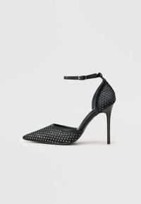 Zapato de tacón alto de malla negra con punta afilada, correa en el tobillo y detalles de tachuelas plateadas en la punta y en la parte del talón.