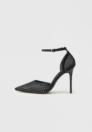 Chaussure à talon haut en maille noire avec bout pointu, bride de cheville et détails en clous argentés sur les zones du bout et du talon.