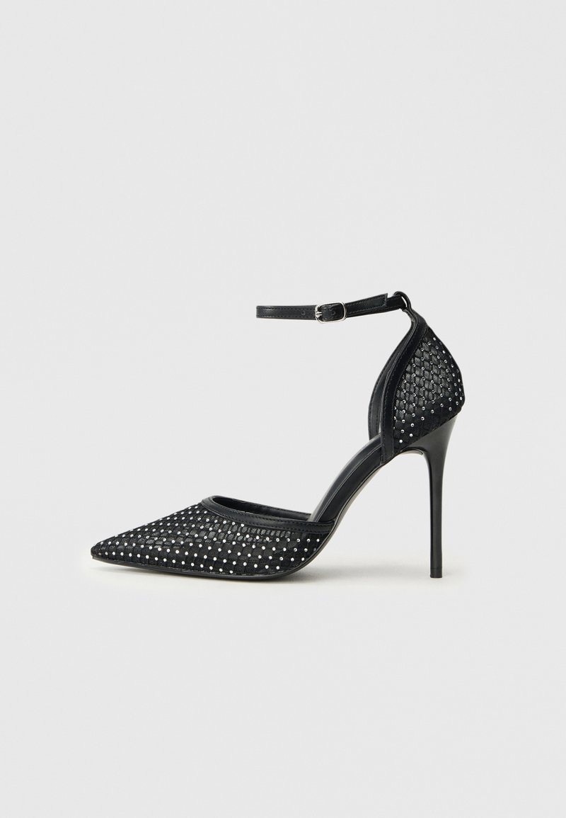 Zapato de tacón alto de malla negra con punta afilada, correa en el tobillo y detalles de tachuelas plateadas en la punta y en la parte del talón.