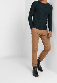 Mörk teal texturerad stickad tröja, beige slim-fit chinos och svarta läder anklarstövlar. Chinosen uppvikta vid fållarna, minimalistisk design.
