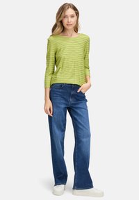 Grünes Langarmoberteil mit strukturierten Wellenmustern, kombiniert mit blauen weiten Jeans und weißen Sneakers, vor einem schlichten Hintergrund stehend.