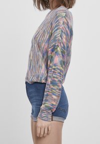 Donna che indossa un maglione lavorato a maglia multicolore con maniche lunghe e pantaloni corti di jeans arrotolati, in piedi di profilo su uno sfondo semplice.
