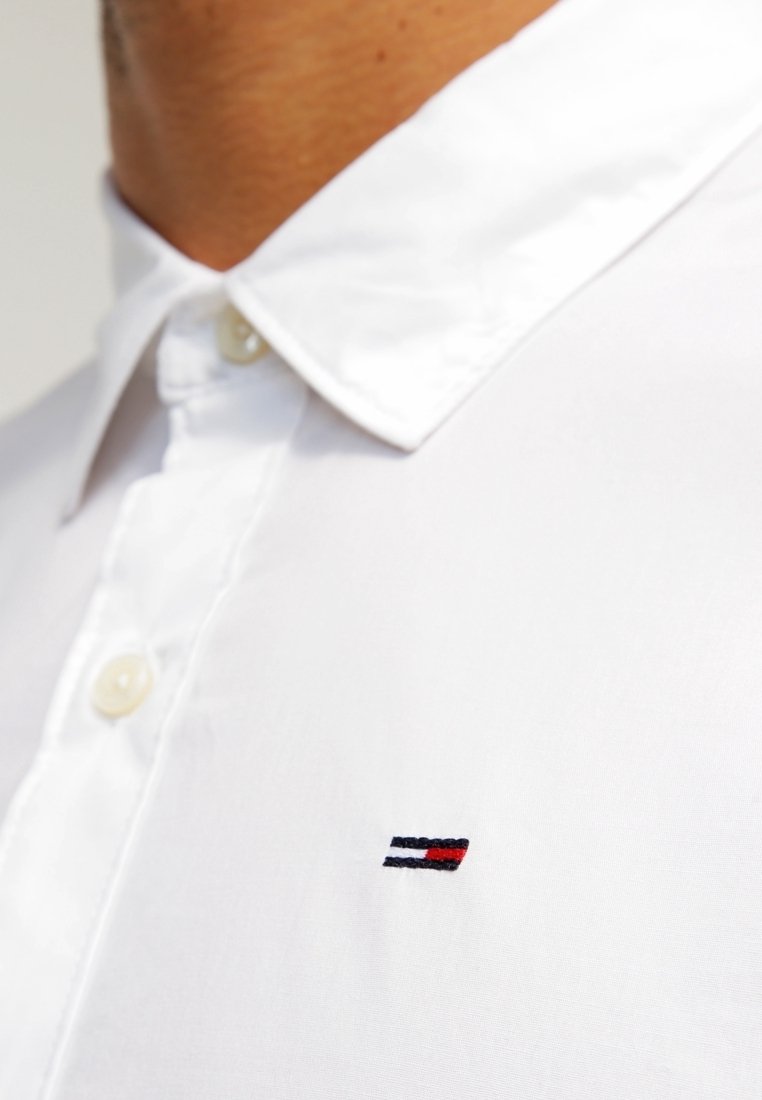 Gros plan d'une personne portant une chemise blanche boutonnée avec un petit logo rayé rouge, blanc et bleu sur la poitrine.