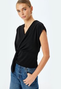 La blusa negra presenta un escote en V, un detalle frontal retorcido y mangas cortas, acompañada de unos pantalones vaqueros de tiro alto en azul claro. Textura suave.