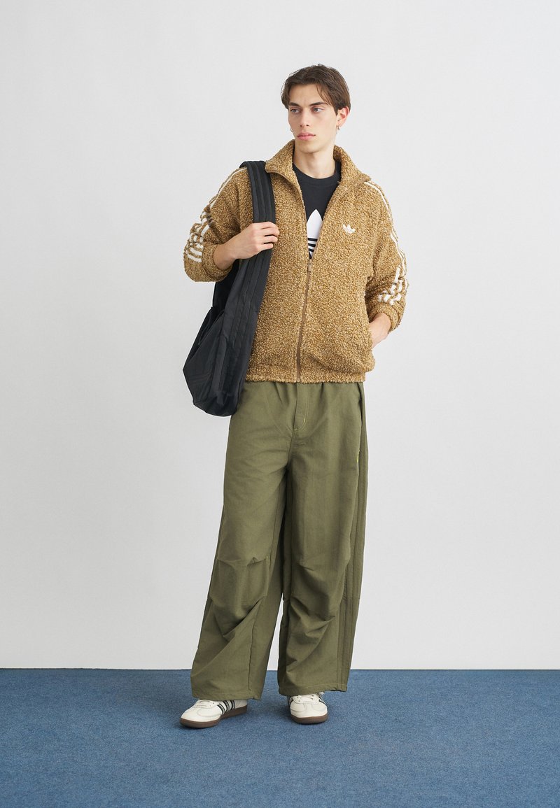 Fuzzy brown zip-up jacket with white stripes, paired with loose olive green pants. Black bag and white sneakers complete the outfit.