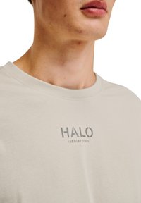 Lys beige bomulds t-shirt med rund halsudskæring og korte ærmer. Har trykt "HALO" logo og numre i mørkegrå på forsiden.