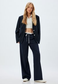 Blazer ajusté marine sur un polo blanc cropped, associé à un pantalon large assorti et des chaussures à enfiler crème. Tissu doux et coupe décontractée.