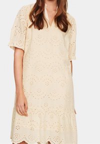 Vaalean beige -värinen mekko, joka on valmistettu tekstuuri-kankaasta ja jossa on neulottuja silmukoita, V-mallinen kaula-aukko, lyhyet puffihihat ja frillahelma.