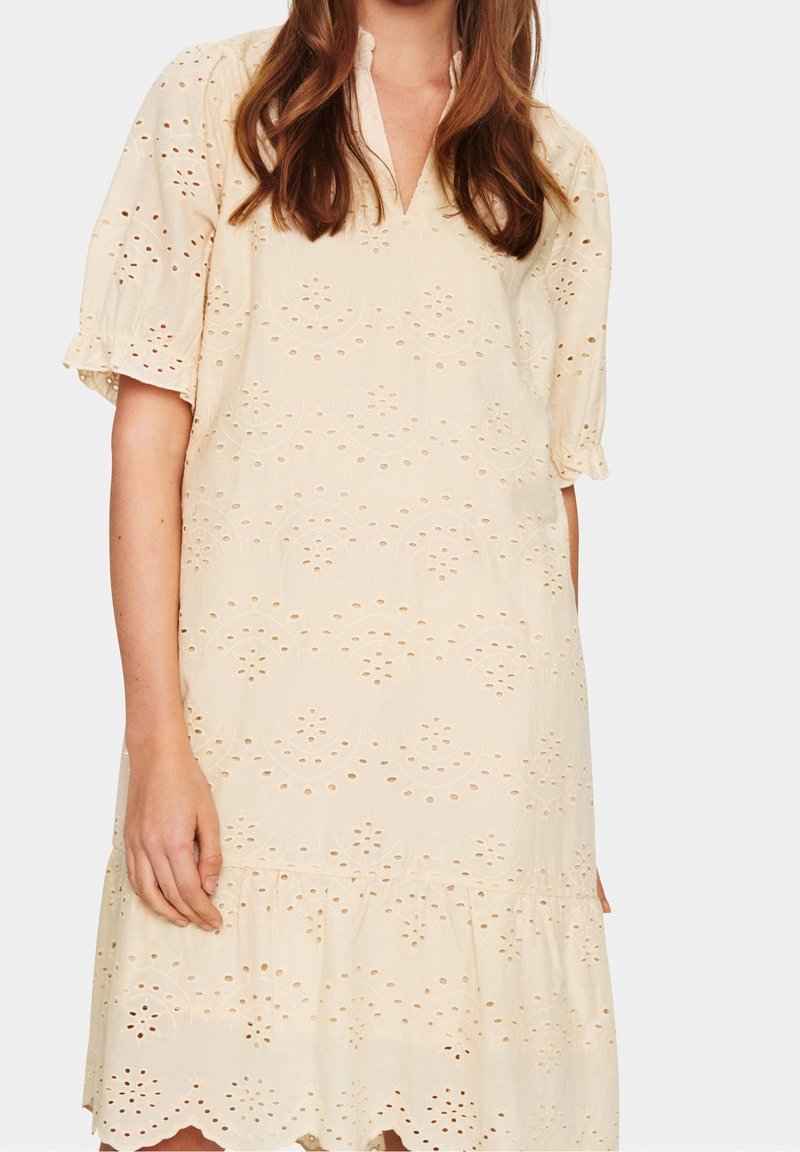 Vaalean beige -värinen mekko, joka on valmistettu tekstuuri-kankaasta ja jossa on neulottuja silmukoita, V-mallinen kaula-aukko, lyhyet puffihihat ja frillahelma.