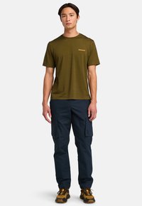Olijfgroene T-shirt met een gestructureerde afwerking en een klein logo, gecombineerd met marineblauwe cargobroek met meerdere zakken en donkerbruine wandelschoenen.