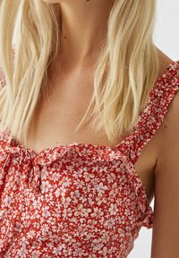 Robe rouge à fleurs comportant de petites fleurs blanches, des bretelles froncées et un décolleté plissé. Tissu léger avec une texture douce.