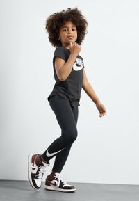 Fekete Nike póló fehér logóval, fekete leggings és fekete, fehér és barna színű, piros részletekkel ellátott magas szárú cipő, álló pózban.