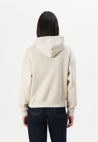 Hoodie beige avec une capuche à cordon de serrage, des poignets et un ourlet côtelés. Porté avec un jean en denim foncé, affichant une coupe décontractée et une texture lisse.