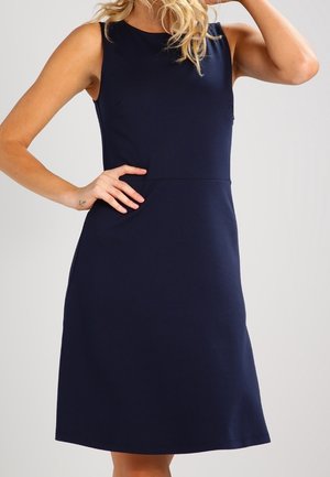 Robe sans manches bleu marine longueur genou portée par une personne avec une main sur la hanche et l'autre main levée près de la tête sur un fond uni.