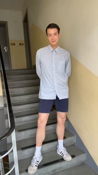 Νεαρό άτομο που στέκεται σε εσωτερικές σκάλες, φοράει ριγέ πουκάμισο, ναυτικό σορτς, λευκές κάλτσες και λευκά αθλητικά παπούτσια Adidas.