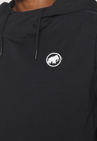 Sudadera negra de tela suave, que presenta un logo de mamut blanco dentro de un contorno circular en el lado izquierdo del pecho. Cordón ajustable en la capucha.