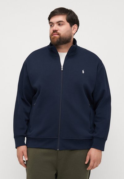 Marineblauwe rits-sweater gemaakt van zachte stof, met een hoge kraag, zijzakken en een klein wit logo op de borst.