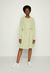 Vestido verde-claro com botão, de mangas compridas e cintura ajustável, combinado com ténis brancos. Textura suave, design até aos joelhos.