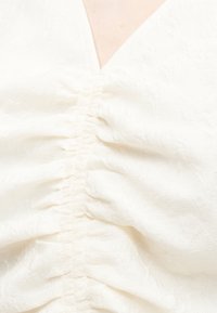 Gros plan sur un tissu blanc texturé froncé avec un motif floral subtil et une bordure d'encolure en V visible.