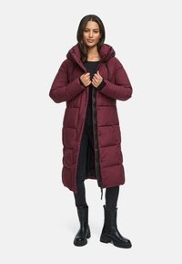 Cappotto lungo imbottito bordeaux con cappuccio, dotato di chiusura con zip e bottoni a pressione, design trapuntato e vestibilità rilassata. Indossato con jeans neri e stivali.