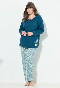 Kvinde iført teal langærmet top med blomsterdetaljer og lyseblå stribede blomsterpyjamasbukser, stående barfodet mod hvid baggrund.