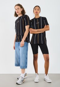 Svarta t-shirts med vertikala orange och vita ränder, kombinerade med denimshorts och svarta cykelbyxor. Båda bär sneakers.