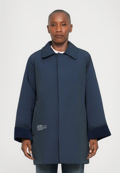 Cappotto blu navy con colletto strutturato, maniche oversize e polsini testurizzati. Presenta dettagli testuali bianchi sul davanti in basso.