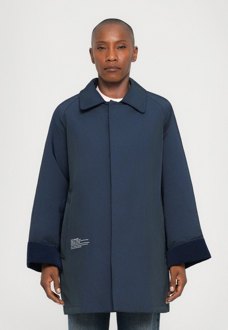 Navy blue coat with a structured collar, oversized sleeves, and textured cuffs. Features white text detailing on the lower front.