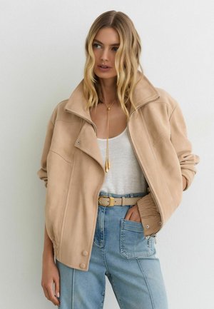 Blonde Frau trägt eine lockere beige Jacke, weißes Tanktop, blaue Jeans mit großen Vordertaschen, einen beigen Gürtel und eine lange goldene Halskette vor weißem Hintergrund.