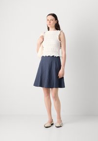 White textured sleeveless top with ruffled edges, paired with a dark blue A-line skirt. Worn with light-colored flat shoes.