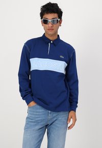 Morska dolga majica polo z lahkotnim modrim vodoravnim pasom, na katerem je napis "LACOSTE" ter majhnim zelenim krokodilom na prsih.