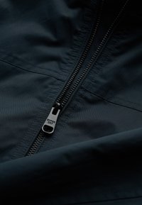 El tejido de la chaqueta azul oscura presenta una cremallera negra con la marca "SUPER DRY", una textura suave y un detalle de costura a lo largo de las costuras.