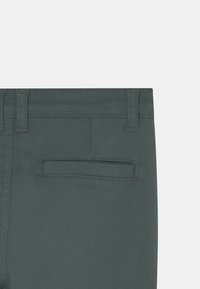 Tmavě zelené kalhoty z hladkého bavlněného směsového materiálu, s poutky na opasek a zadní kapsou s vodorovným otvorem. Viditelné švy.