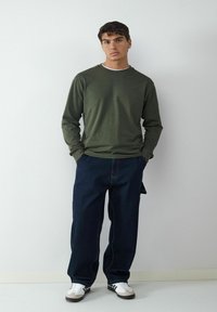 Chemise à manches longues vert olive avec un col rond, assortie à un jean en denim bleu foncé et des baskets blanches avec des accents noirs.