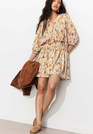 Femme portant une robe beige à motifs floraux avec des manches bouffantes et une taille smockée, tenant une veste marron, debout contre un mur blanc.