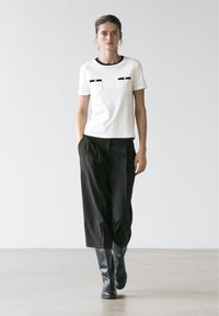 Femme marchant vêtue d'un pantalon noir large et court, d'un haut blanc à manches courtes avec des accents noirs, et de bottes noires montantes, sur un fond uni.